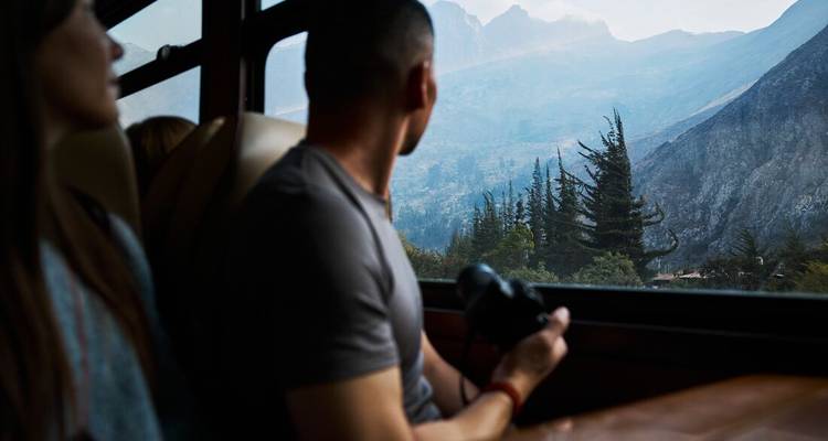 A traveller inside a train gazes out the window toward misty Andean peaks while holding a camera.