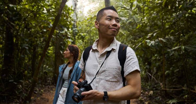 Two trekkers explore dense Amazon jungle, one holding a camera ready for wildlife shots.