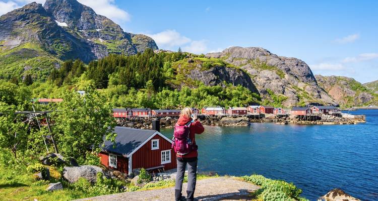 Un routard photographie des rangées de cabanes de pêcheurs rouges à côté d'un fjord bleu vif sous des pentes vertes.