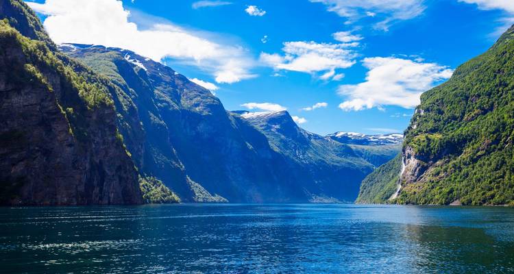 Un fjord bleu profond flanqué de falaises escarpées et boisées et de sommets enneigés s'étend sous un ciel vibrant.
