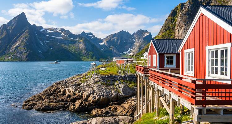 Les cabines rouges emblématiques rorbuer sur pilotis se dressent à côté d'un fjord turquoise dominé par les montagnes déchiquetées et dramatiques des Lofoten sous un ciel bleu.