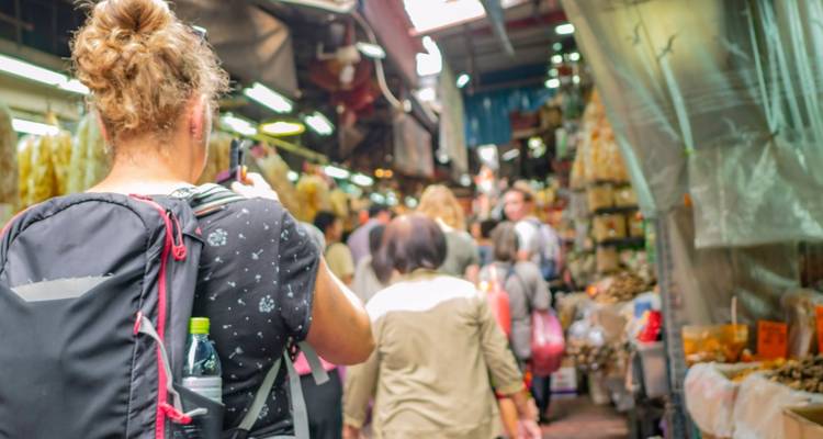Vue par-dessus l'épaule d'un touriste en randonnée photographiant une allée de marché intérieur étroite et bondée.