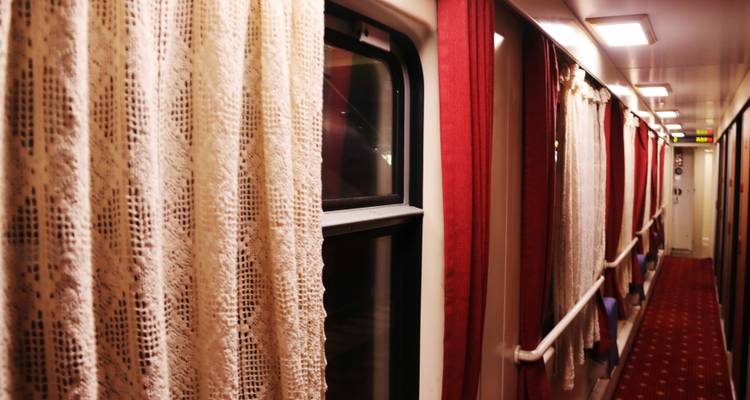Narrow train corridor with lace curtains and red carpet running alongside sleeping cabins.