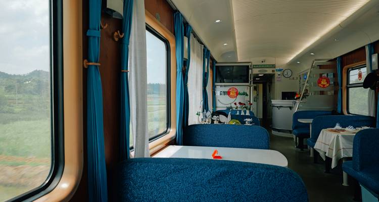 Bright dining-lounge car with blue upholstered booths, white curtains and scenic views out the windows.