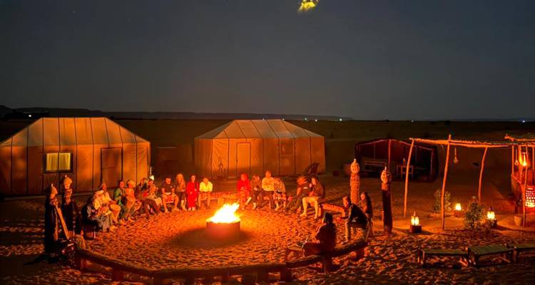 Een grote groep reizigers geniet van een kampvuur buiten woestijntenten onder de sterrenhemel.