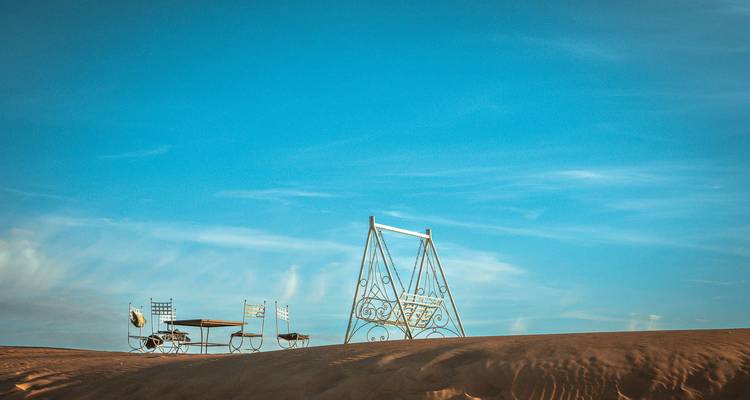 Minimalistische scene van een metalen schommel en klein cafeetje geïsoleerd op een gladde zandduinen onder een enorme blauwe lucht.