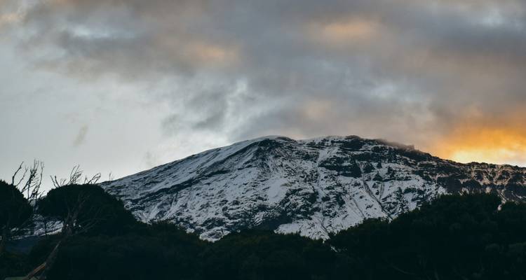 Met sneeuw bedekte bergrug van Kilimanjaro afgetekend tegen dramatische oranje en grijze zonsondergangswolken.