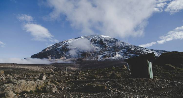 Steile, sneeuwgestreepte wand van de Kilimanjaro torens uit boven rotsachtige grond en een klein houten hutje.