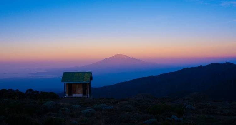 Pastelkleurige dageraadkleuren vervagen boven de verre Mount Meru voorbij een klein huisje dat op het plateau balanceert.