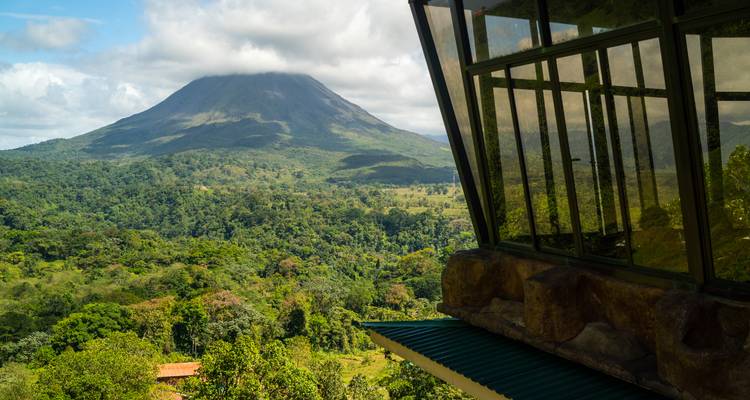 Panoramisch uitzicht op de Arenal-vulkaan omlijst door de glazen ramen van een lodge op een helling.