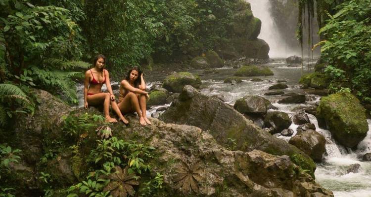 Two young women in swimsuits sit on mossy rocks beside a lush jungle river with a tall waterfall in the background