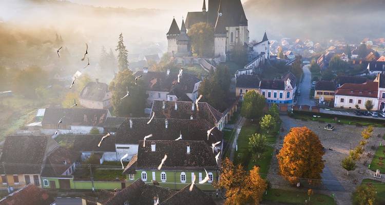 Misty sunrise aerial of a fortified Transylvanian church rising above tiled rooftops and autumn trees.