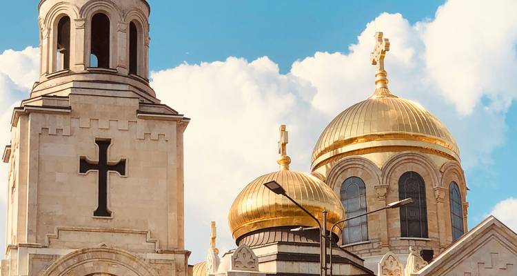 Close-up of golden domes and stone bell tower of Varna Cathedral against fluffy clouds.