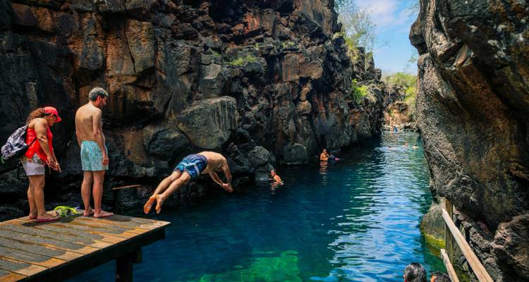 Des aventuriers se lancent d'une plateforme en bois dans une piscine d'un canyon volcanique aux eaux cristallines tandis que d'autres observent depuis des rebords rocheux.