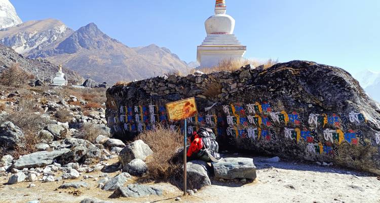 Mur de mani rocheux peint d'inscriptions tibétaines à côté de stupas blancs sur un sentier de montagne.