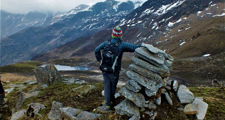 Un randonneur se tient près d'un cairn et contemple un terrain alpin accidenté parsemé de plaques de neige.