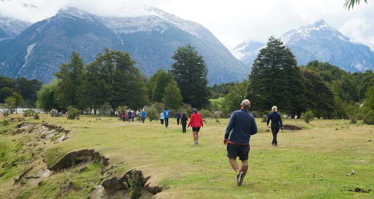 Walkers cross a grassy riverside plain framed by dramatic Patagonian mountains.