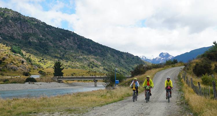 Three cyclists pedal a gravel riverside road beneath green hills and distant snowy summits.
