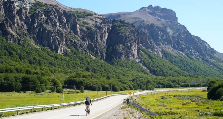 Cyclist travels a scenic road flanked by steep green cliffs and forested slopes.