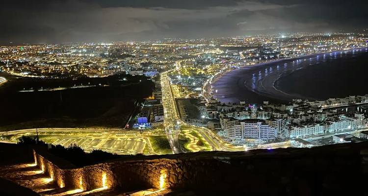 Panorama nocturne d'une ville côtière avec routes du littoral illuminées et reflets sur la baie.