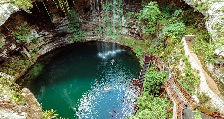 Aerial view of a turquoise cenote with waterfall and swimmers surrounded by lush limestone walls