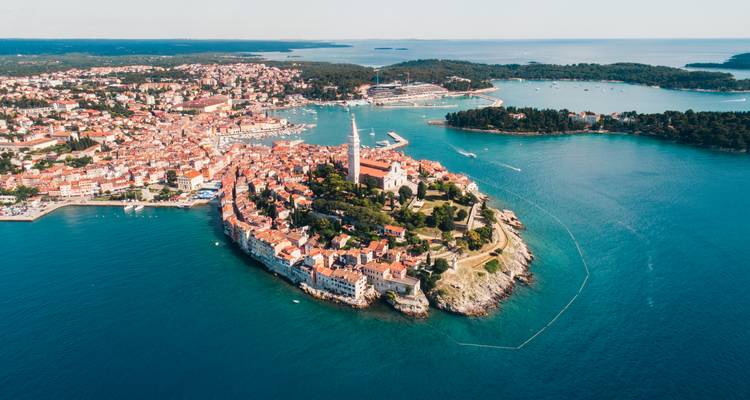 Luftaufnahme einer historischen Hafenstadthalbinsel mit einem hohen Kirchturm, umgeben von türkisem Meer.