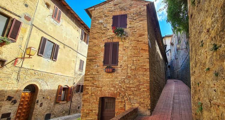 Un callejón estrecho de ladrillos en un pueblo medieval toscano con casas de piedra, persianas de madera y cestas de flores colgantes bajo un cielo azul brillante.