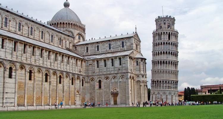 La Torre Inclinada de Pisa y la Catedral de Pisa de pie una al lado de la otra en un prado verde con turistas reunidos en la base en un día nublado.