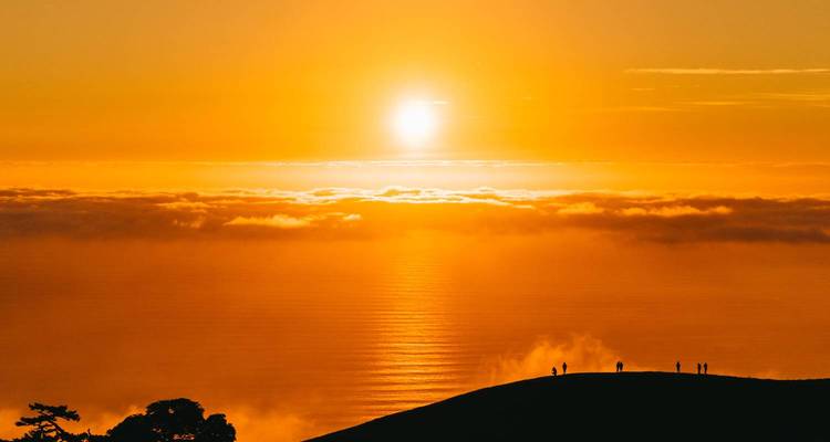 Brilliant orange sunset above a sea of clouds with small silhouette hikers on a ridge.