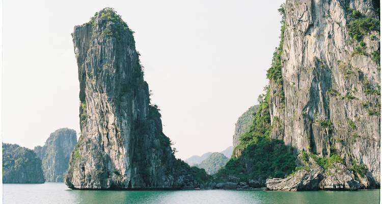 Tall narrow limestone pillar towers over emerald water between two larger karst formations in Ha Long Bay