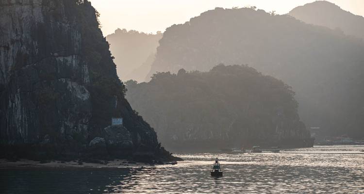 Sunlit haze silhouettes jagged islands while a small buoy floats on shimmering water in Ha Long Bay