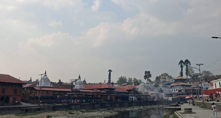 Smoke rises from funeral ghats along a wide sacred river at Pashupatinath with temples silhouetted against a hazy sky.
