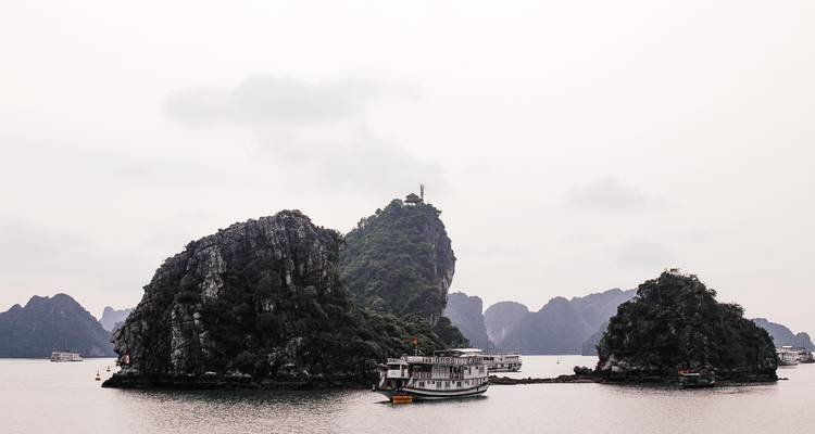 Traditional junk boat sails between small jungle-covered islands under a hazy sky in Ha Long Bay.
