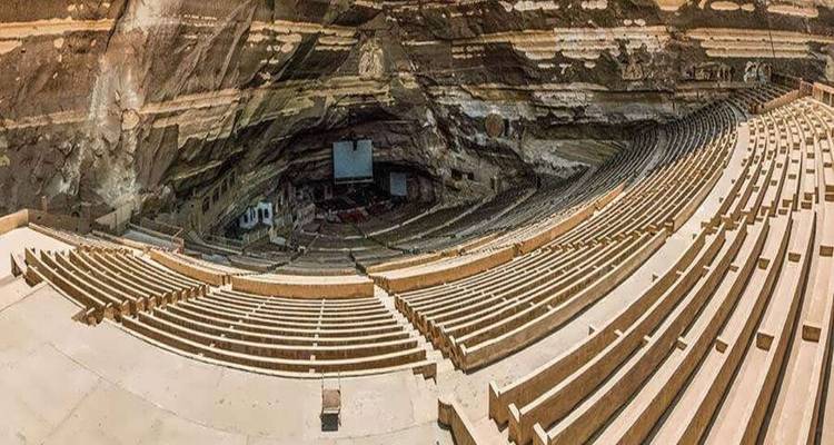 Expansive panoramic view of an enormous rock-hewn amphitheatre with tiered stone seating curving around a stage area.
