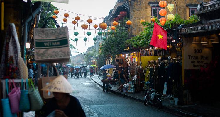 Rain-soaked lantern street in Hoi An old town with shoppers under umbrellas and Vietnamese flag