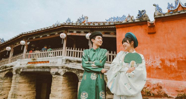 A young couple in traditional Vietnamese ao dai pose smiling in front of Hoi An’s Japanese Covered Bridge.