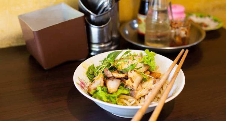 A bowl of Vietnamese noodles topped with grilled pork, herbs, and crispy toppings sits on a dark wooden table.
