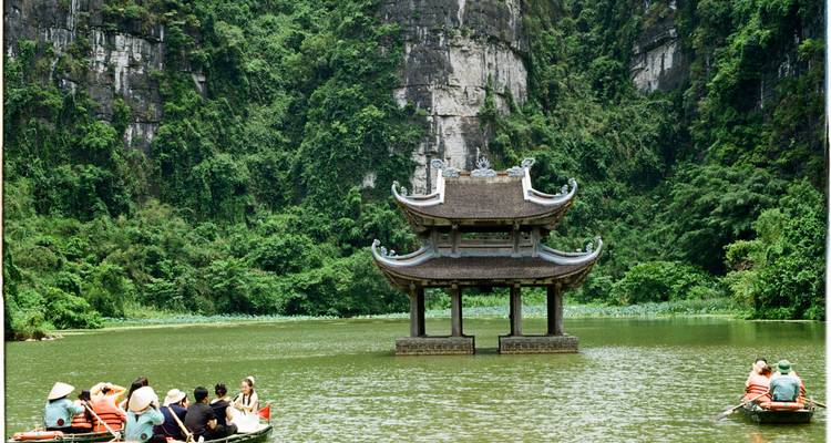 Boats with tourists paddling on a river toward a small pagoda nestled in karst cliffs in Ninh Binh