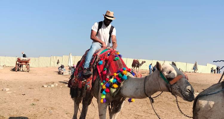 Un voyageur coiffé d'un chapeau chevauche un chameau drapé de couvertures colorées à travers un enclos sablonneux par une journée dégagée.