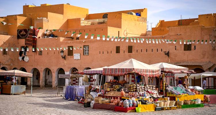Bustling desert town square with colorful market stalls against ochre adobe buildings under bright skies.