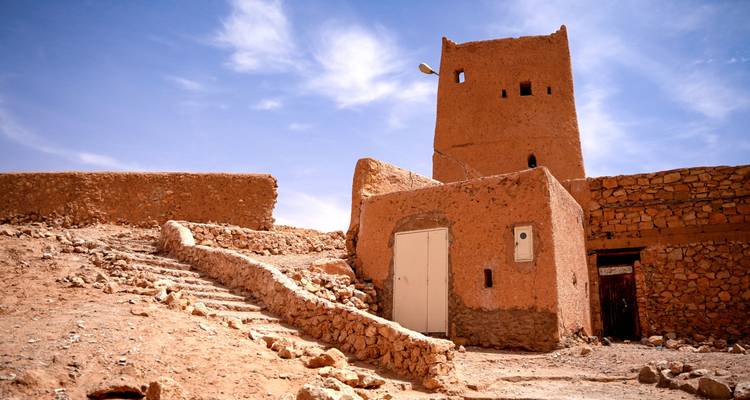 Earthen desert tower and surrounding adobe walls perched on a rocky hill under a cloud-dotted sky.