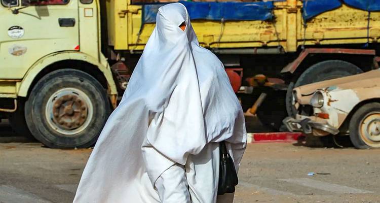 Woman wrapped head-to-toe in a flowing white traditional garment walks along a dusty urban street.