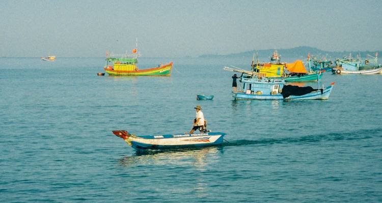 A fisherman stands in a small wooden boat among colorful fishing vessels on calm blue water