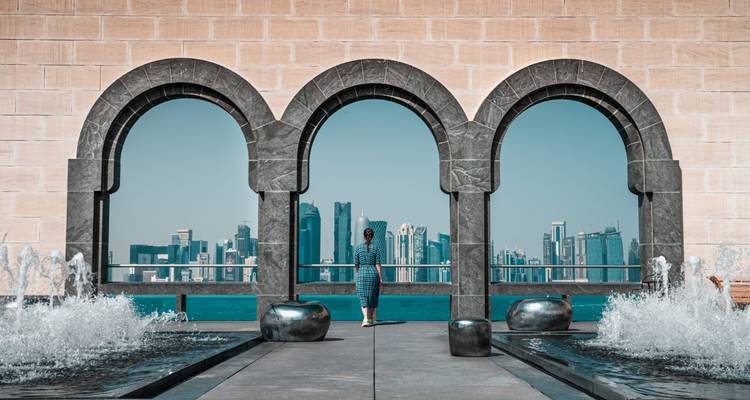 Femme se tenant entre trois grandes arcades en pierre du Musée de l'Art Islamique, regardant vers l'horizon urbain moderne de Doha de l'autre côté d'une étendue d'eau bleue et de fontaines.