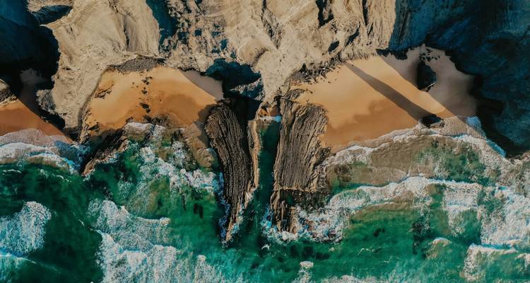 Vue aérienne d'une côte rocheuse spectaculaire avec des vagues bleu-vert et des petites baies de sable séparées par des crêtes de pierre noire.