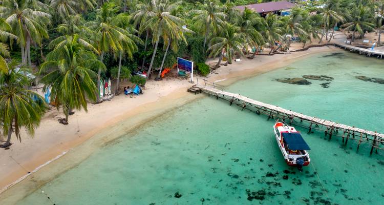 Aerial view of a quiet tropical cove with pale sand, palm trees, a long wooden jetty and a small tour boat on emerald water.