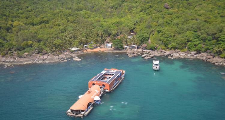 Floating orange-roofed structure and a white yacht moored in clear blue-green water off a densely forested shoreline.