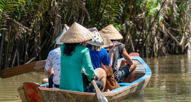 Tourists wearing conical hats paddle a narrow wooden boat through muddy creek surrounded by dense mangrove roots.