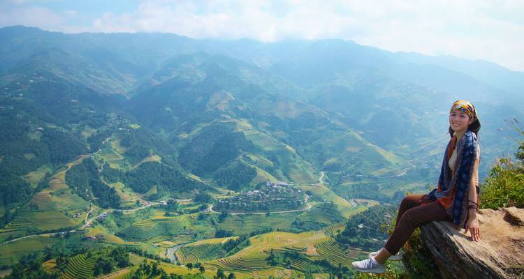 Young woman seated on a rock ledge overlooking expansive green terraced mountains and misty horizons.