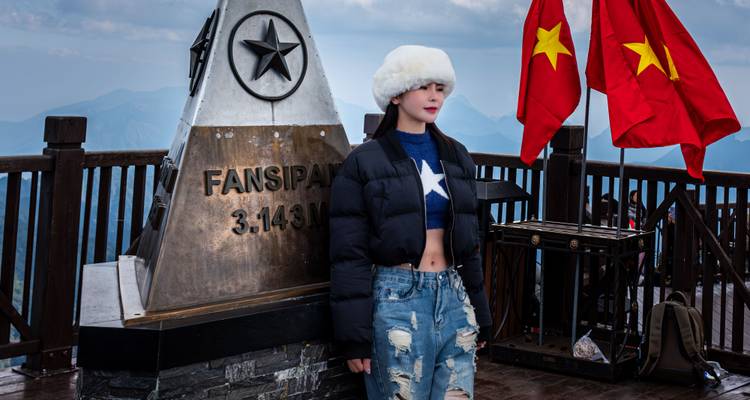 Young woman in winter clothes poses beside the Fansipan summit marker with Vietnamese flags fluttering.